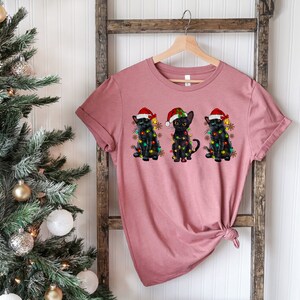 Pode incluir: Uma camiseta rosa com tr&ecirc;s gatos pretos usando chap&eacute;us de Papai Noel e luzes de Natal. Os gatos est&atilde;o dispostos em fila, com o gato &agrave; esquerda usando um chap&eacute;u de Papai Noel vermelho, o gato do meio usando um chap&eacute;u de Papai Noel verde e o gato &agrave; direita usando um chap&eacute;u de Papai Noel azul.