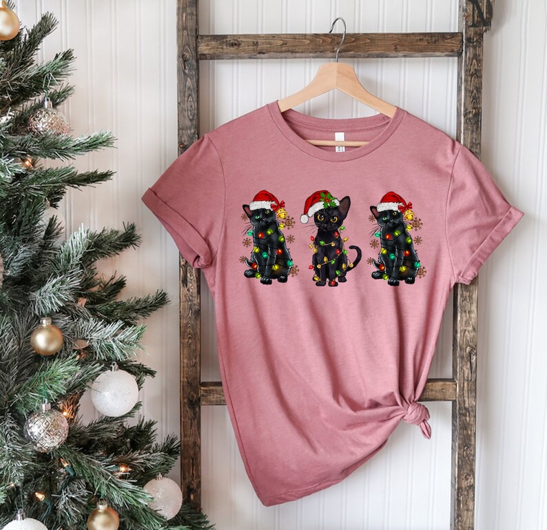 Pode incluir: Uma camiseta rosa com tr&ecirc;s gatos pretos usando chap&eacute;us de Papai Noel e luzes de Natal. Os gatos est&atilde;o dispostos em fila, com o gato &agrave; esquerda usando um chap&eacute;u de Papai Noel vermelho, o gato do meio usando um chap&eacute;u de Papai Noel verde e o gato &agrave; direita usando um chap&eacute;u de Papai Noel azul.