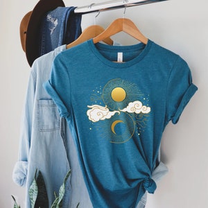 May include: A teal blue t-shirt with a gold sun and moon design. The design features a crescent moon, a sun with rays, and clouds.