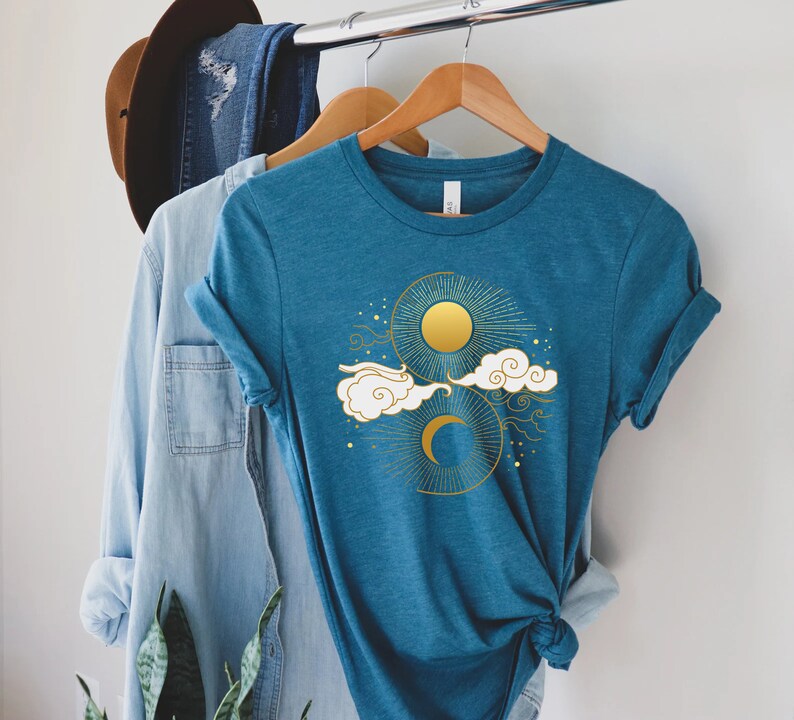 May include: A teal blue t-shirt with a gold sun and moon design. The design features a crescent moon, a sun with rays, and clouds.