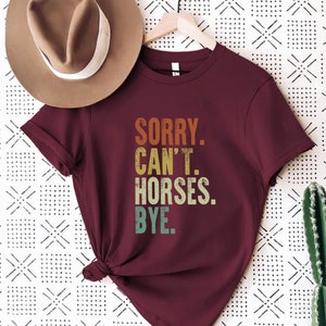 May include: A maroon t-shirt with the text "SORRY. CAN'T. HORSES. BYE." in a vintage style font with a gradient of orange, yellow, green, and blue.