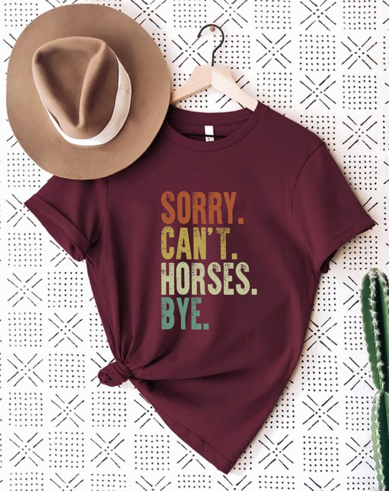 May include: A maroon t-shirt with the text "SORRY. CAN'T. HORSES. BYE." in a vintage style font with a gradient of orange, yellow, green, and blue.