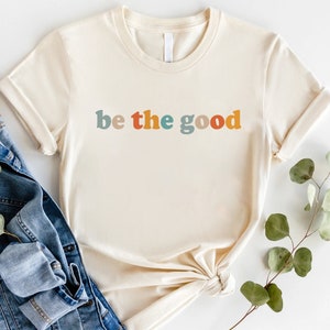 May include: Cream-colored t-shirt with the phrase "be the good" in colorful, retro-style lettering. The shirt has short sleeves and a crew neck. A denim jacket and greenery are in the background.