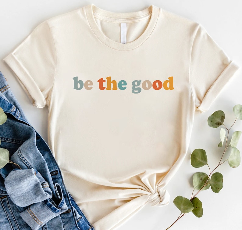 May include: Cream-colored t-shirt with the phrase "be the good" in colorful, retro-style lettering. The shirt has short sleeves and a crew neck. A denim jacket and greenery are in the background.