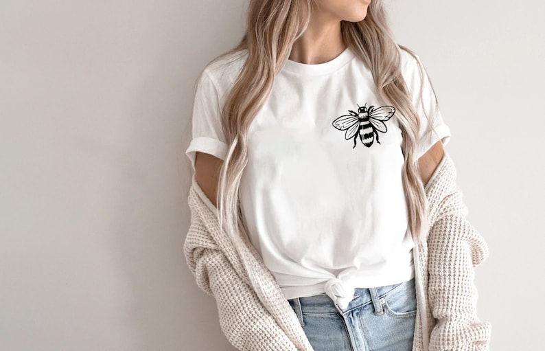 May include: A white short-sleeved t-shirt featuring a black line art graphic of a bee on the upper left chest area. The shirt has a crew neckline and is styled with a knot at the bottom hem, paired with blue denim jeans and a beige textured cardigan.
