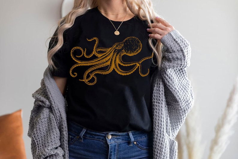 May include: Black t-shirt featuring a detailed gold octopus illustration. The octopus design is centered on the shirt, showcasing its tentacles and body. The shirt is a classic crew neck style.