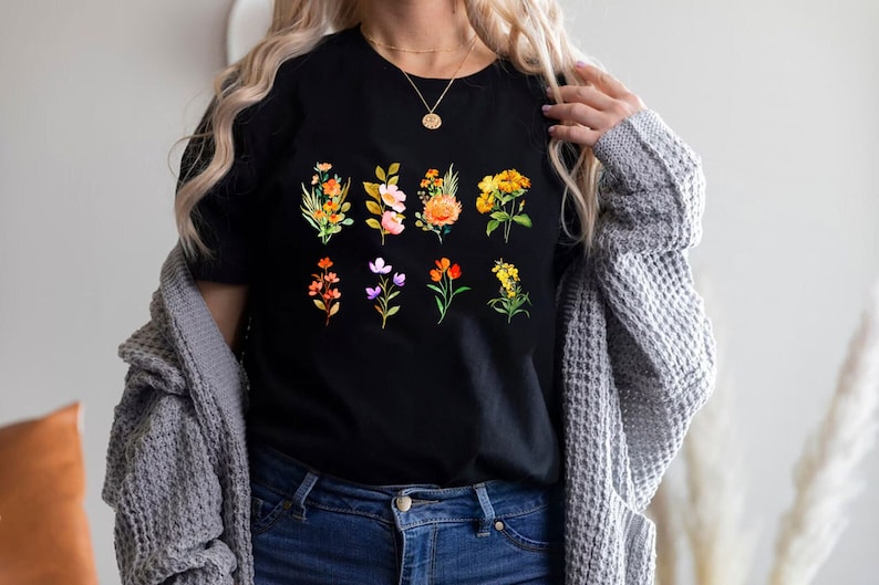 Może przedstawiać: Czarny t-shirt z nadrukiem kwiatowym przedstawiającym r&oacute;żne kwiaty w odcieniach pomarańczowego, ż&oacute;łtego, r&oacute;żowego i fioletowego.