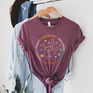 May include: Burgundy t-shirt with a circle graphic featuring the American Sign Language symbol for "I love you" surrounded by the words "Accept", "Adapt", "Advocate", "Equality", "Inclusion", and "Unity".