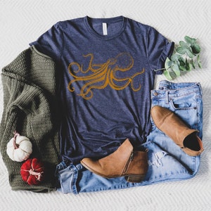 May include: Navy blue t-shirt with an orange octopus design. The shirt is paired with blue jeans, brown boots, and a green sweater. Decorative pumpkins and eucalyptus sprigs complete the casual ensemble.
