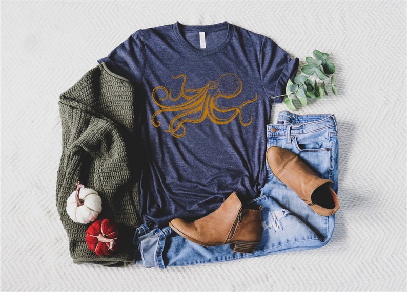 May include: Navy blue t-shirt with an orange octopus design. The shirt is paired with blue jeans, brown boots, and a green sweater. Decorative pumpkins and eucalyptus sprigs complete the casual ensemble.
