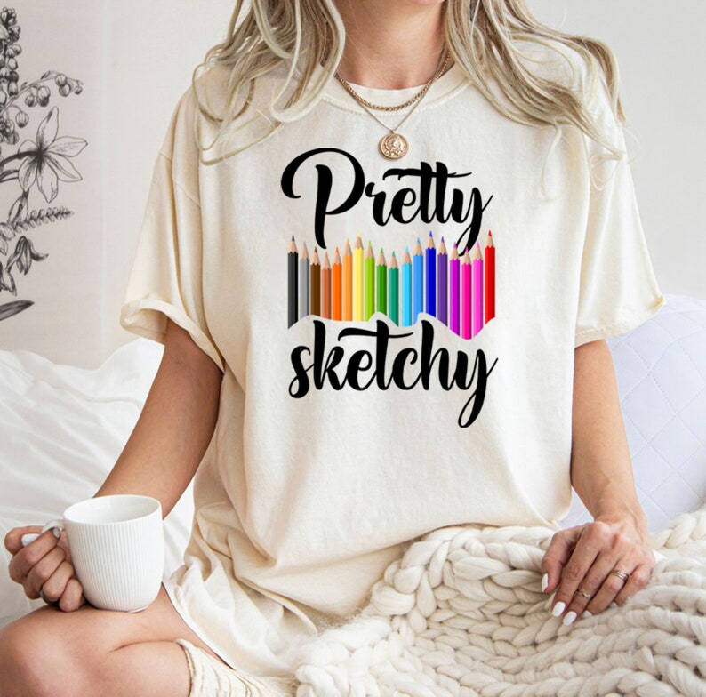 以下が含まれることがあります： 白いTシャツに黒い文字で「Pretty sketchy」と書かれています。テキストは、カラフルな鉛筆の列で囲まれています。