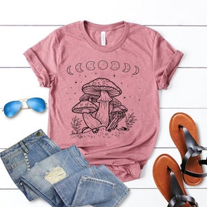 May include: A pink t-shirt with a black and white graphic of mushrooms and moons. The graphic includes the text "Shroom Queens".