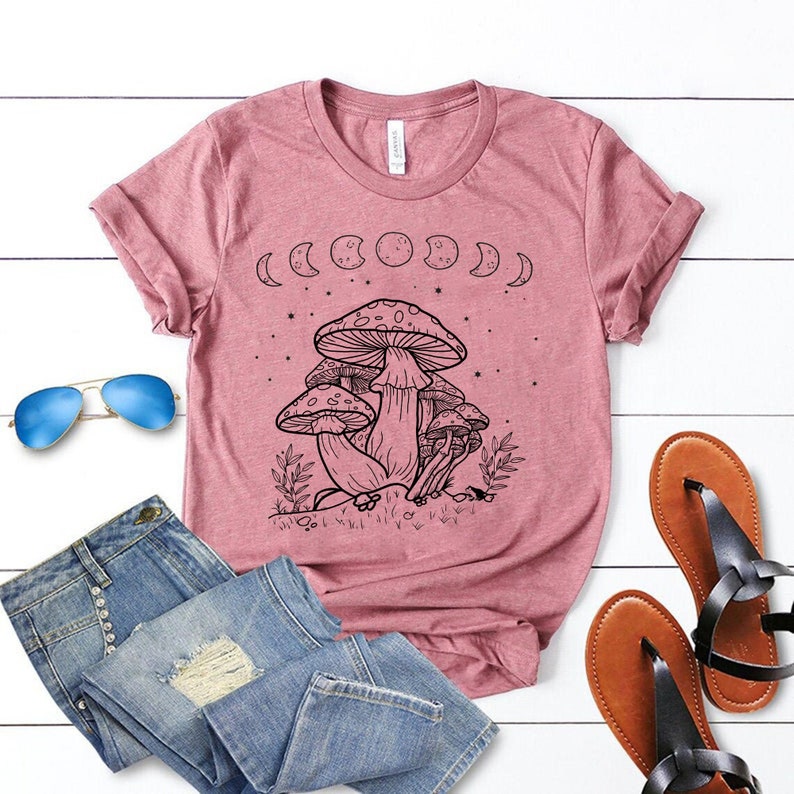 May include: A pink t-shirt with a black and white graphic of mushrooms and moons. The graphic includes the text "Shroom Queens".