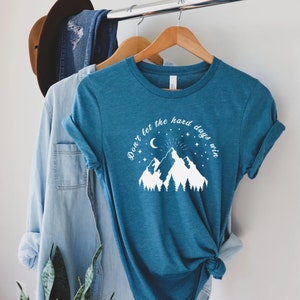 以下が含まれることがあります： 「Don't let the hard days win」の文字が入った、ティール色のTシャツ。デザインには三日月、星、木々のある山脈が含まれています。木製のハンガーにかけられています。