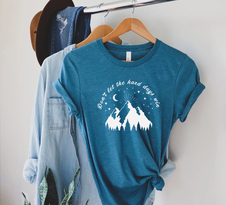 以下が含まれることがあります： 「Don't let the hard days win」の文字が入った、ティール色のTシャツ。デザインには三日月、星、木々のある山脈が含まれています。木製のハンガーにかけられています。