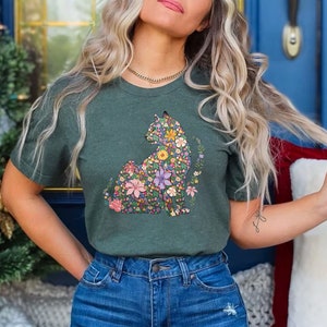 May include: A dark green t-shirt featuring a floral cat design. The cat is composed of colorful flowers in shades of pink, purple, yellow, and blue. The shirt is paired with blue denim jeans. The background includes a Christmas tree and a blue door.