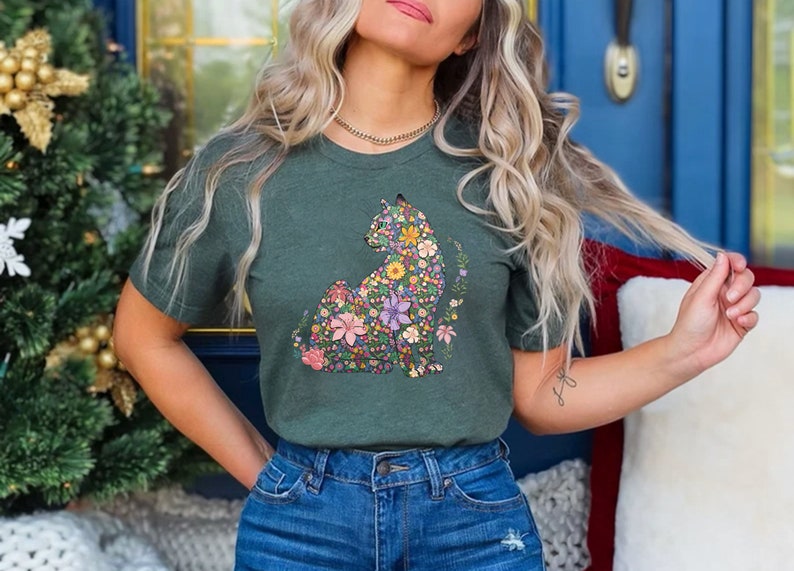 May include: A dark green t-shirt featuring a floral cat design. The cat is composed of colorful flowers in shades of pink, purple, yellow, and blue. The shirt is paired with blue denim jeans. The background includes a Christmas tree and a blue door.