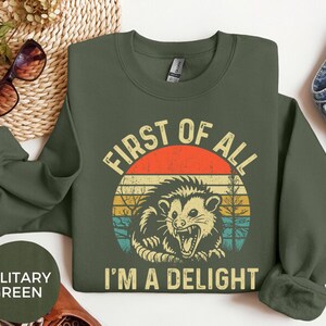 May include: Military green sweatshirt with a retro graphic of an opossum inside a circle with the text "FIRST OF ALL I'M A DELIGHT". The design features a red and orange sun with yellow, blue, and green stripes.