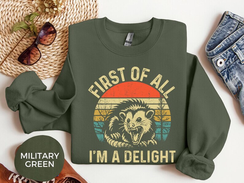 May include: Military green sweatshirt with a retro graphic of an opossum inside a circle with the text "FIRST OF ALL I'M A DELIGHT". The design features a red and orange sun with yellow, blue, and green stripes.
