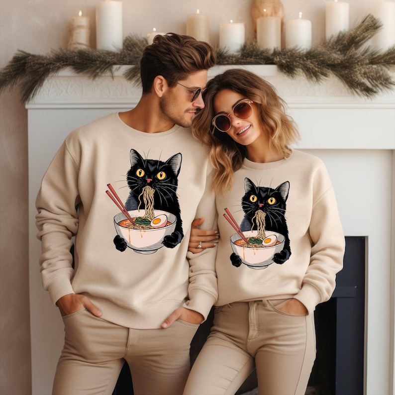 May include: A couple wearing matching beige sweatshirts with a black cat eating ramen noodles printed on the front. The cat is holding chopsticks and a bowl of ramen with an egg.