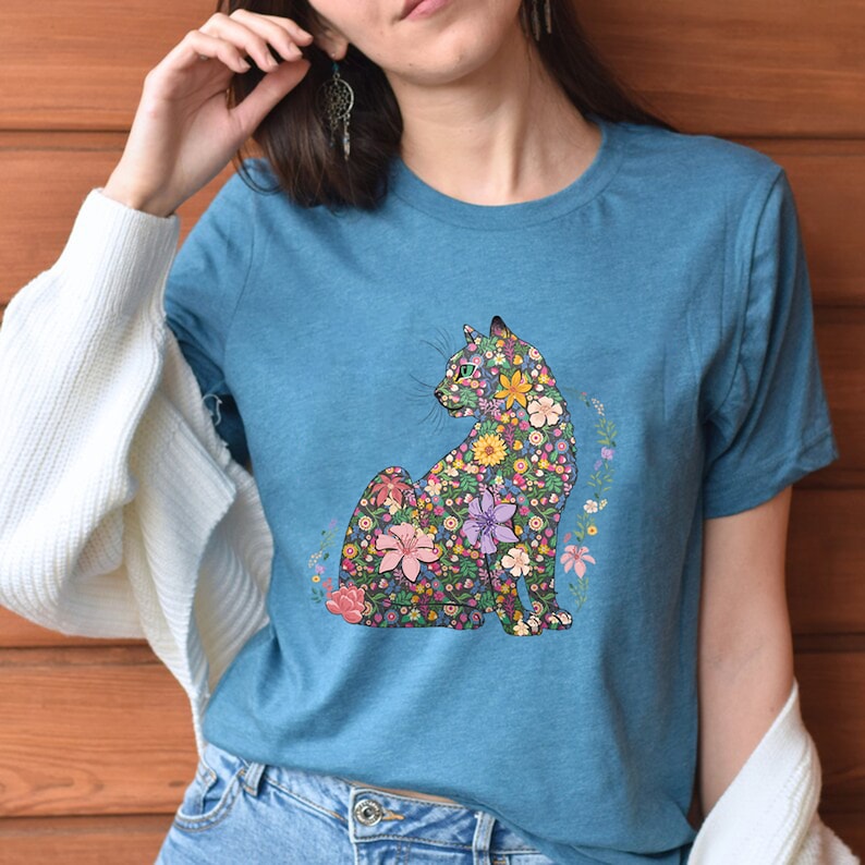 May include: A teal t-shirt featuring a floral cat design. The cat is composed of various colorful flowers, including pink, yellow, and purple blooms. The shirt has short sleeves and a crew neck.