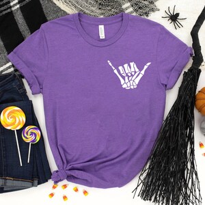 Op de afbeelding: Paars T-shirt met een witte skelet hand die een shaka-teken maakt. Het shirt is omringd door Halloween-decoraties, waaronder een bezem, snoepmais en pompoenen.