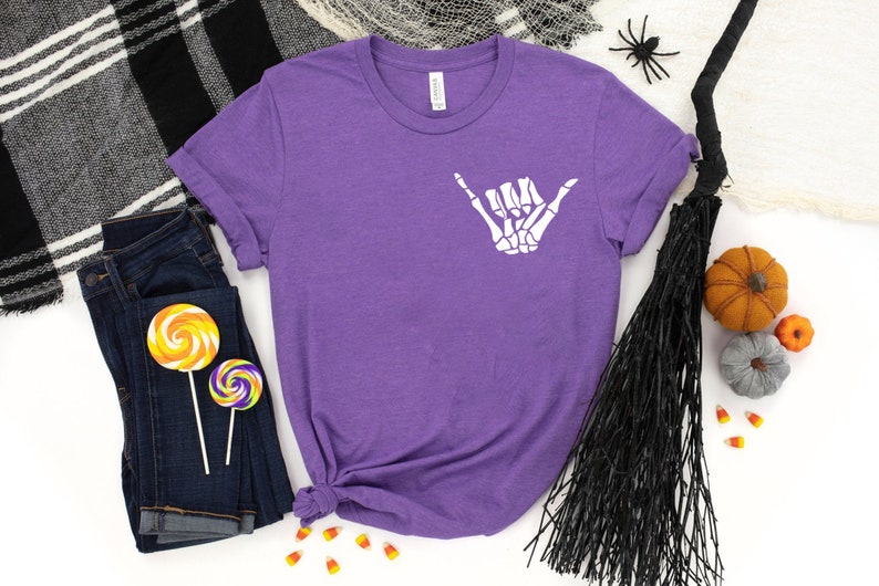 Op de afbeelding: Paars T-shirt met een witte skelet hand die een shaka-teken maakt. Het shirt is omringd door Halloween-decoraties, waaronder een bezem, snoepmais en pompoenen.