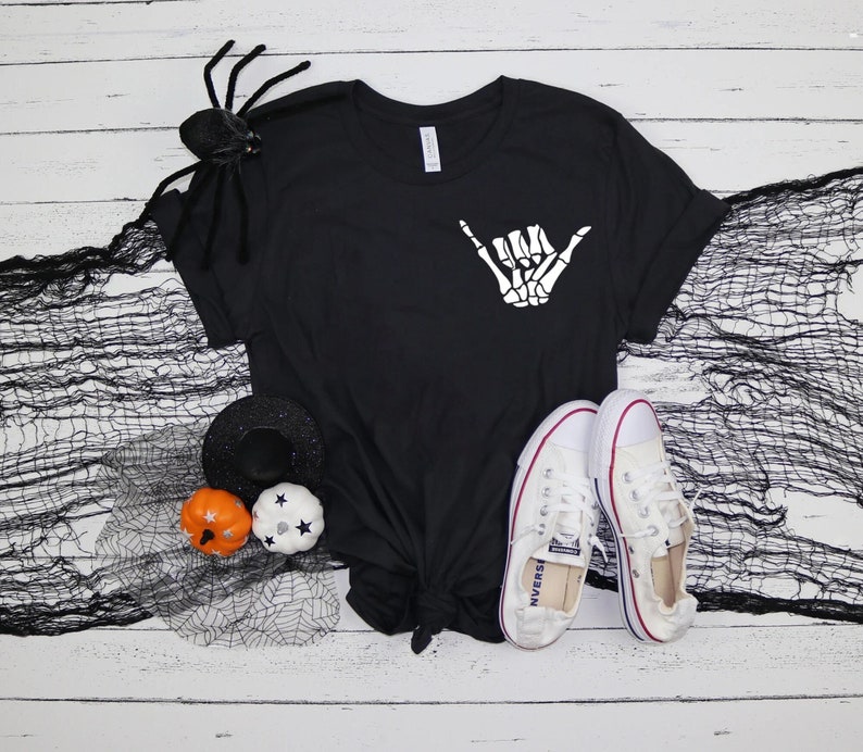 Op de afbeelding: Zwart T-shirt met een witte skelethand die een shaka-teken maakt. Het shirt ligt op een wit houten oppervlak met een zwarte spin, een zwarte heksenhoed en twee kleine pompoenen.