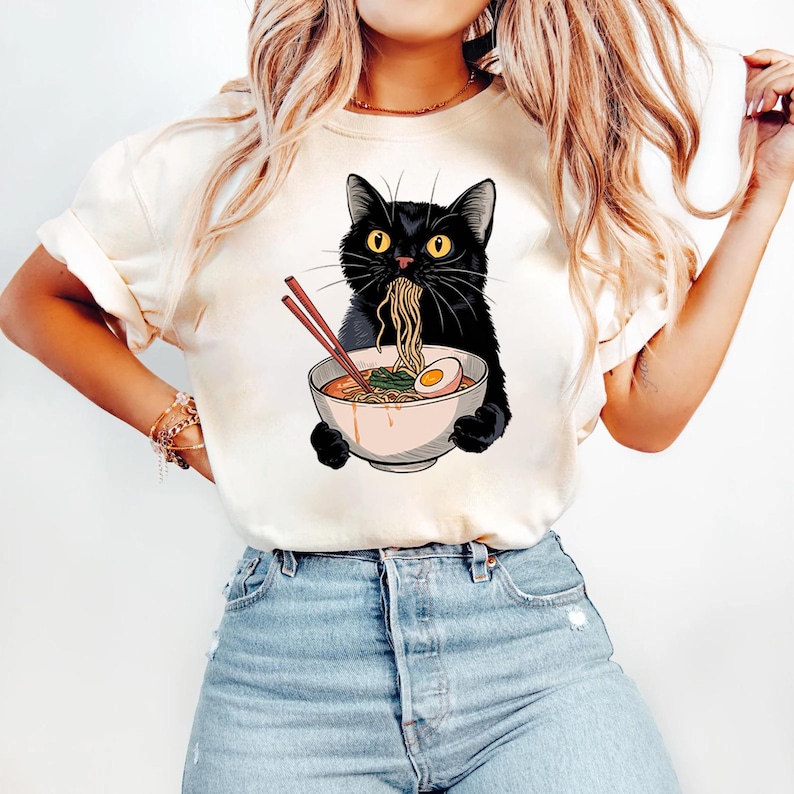 May include: A white t-shirt with a black cat eating ramen noodles from a pink bowl. The cat has yellow eyes and is wearing a red collar. The shirt has a graphic design of the cat eating ramen.