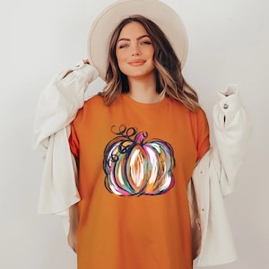 Może przedstawiać: Pomarańczowy t-shirt z centralną grafiką dyni, zilustrowaną za pomocą abstrakcyjnych, wielokolorowych pociągnięć pędzla w odcieniach niebieskiego, r&oacute;żowego, białego i ż&oacute;łtego, w połączeniu z białym kapeluszem z szerokim rondem i białą koszulą.