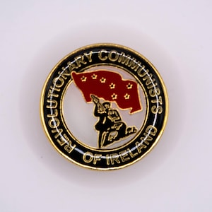 Op de afbeelding: Een gouden speld met een zwarte cirkel rond de rand. De cirkel heeft de woorden "Revolutionary Communists of Ireland" eromheen geschreven. Binnen de cirkel bevindt zich een rode vlag met zeven witte sterren en een zwart silhouet van een persoon die een geweer vasthoudt.