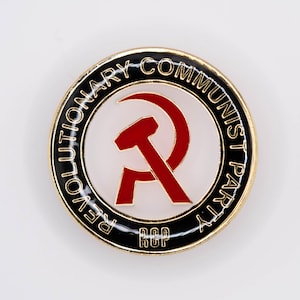 Może przedstawiać: Złota szpilka z czarno-białym kółkiem. Na kółku widnieje napis "Revolutionary Communist Party" wokół krawędzi. Wewnątrz kółka znajduje się czerwony symbol młota i sierpa z literami "RCP" w środku.