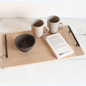 Può includere: Un vassoio di legno con manici in metallo nero, due tazze in ceramica beige, una piccola ciotola nera e un libro bianco con testo nero.