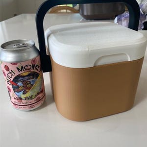 May include: A tan and white cooler with a black handle next to a can of "Bitch Monkey" beer. The beer can is pink with a cartoon monkey and text. The cooler is rectangular and appears to be made of plastic.