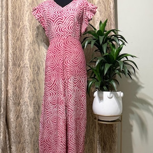 May include: A full-length jumpsuit featuring a vibrant magenta and white geometric wave pattern. The garment has a V-neckline, short flutter sleeves, and a fitted waistline that transitions into wide-leg trousers, displayed on a mannequin.