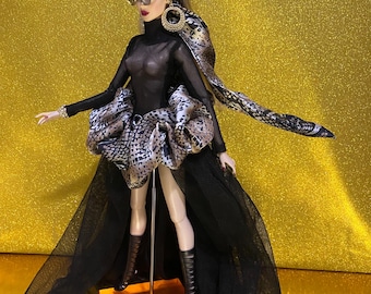 May include: A fashion doll in a black mesh top, skirt, and boots, with a leopard print headscarf and accents. The doll is wearing sunglasses and large gold hoop earrings. The doll is standing on a clear stand.