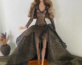 May include: A fashion doll with long blonde hair, wearing a gold and black gown with a flowing train. The dress features a plunging neckline and long sleeves. The doll is wearing gold heels and stands on an orange base.