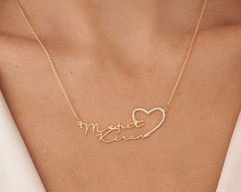 May include: A gold necklace with two names, "Margot" and "Kiran", written in cursive script. The names are connected by a heart-shaped pendant with small diamonds around the edge.