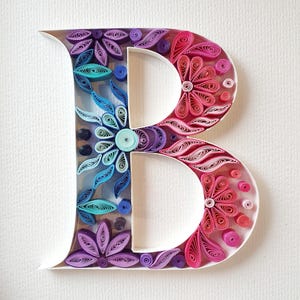 Può includere: Un'opera d'arte di quilling di carta fatta a mano a forma di lettera "B". La lettera è decorata con disegni floreali nei toni del blu, viola e rosa. La carta è arrotolata e modellata per creare un effetto 3D.