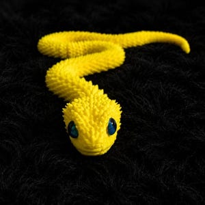 Puede incluir: Una figura de serpiente articulada de color amarillo brillante con una superficie muy texturizada y puntiaguda, y dos ojos azules brillantes y prominentes. La serpiente se muestra en una posición curva y sinuosa, exhibiendo una estructura corporal segmentada y detallada.