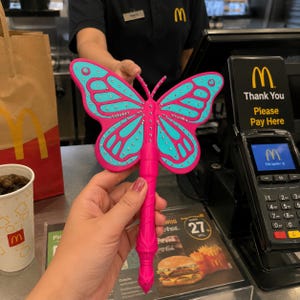 May include: A bright pink plastic butterfly-shaped wand with a handle and turquoise wing accents. The wings are decorated with small, clear embedded rhinestones arranged in lines along the wing veins and edges, creating a sparkling effect.