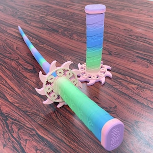 May include: Two toy swords featuring a gradient color scheme transitioning from pink to purple, blue, and green. Each sword has a handle wrapped in a textured pattern and a circular guard designed with small, interlocking gear-like shapes in light pink.