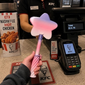 Pode incluir: Uma varinha em forma de estrela com um esquema de cores degradê que transita do roxo no topo para o rosa na base. A varinha possui uma cabeça de estrela de cinco pontas lisa e um cabo texturizado em espiral, segurada num restaurante fast food.