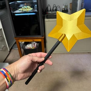 May include: A star-shaped wand featuring a bright yellow, five-pointed star head with a faceted, geometric surface texture. The star is securely attached to a long, smooth, matte black cylindrical handle, creating a distinct contrast between the vibrant yellow top and the dark, neutral base.