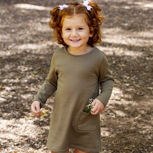 Può includere: Una bambina con i capelli rossi indossa un vestito verde oliva a maniche lunghe con una tasca. Tiene un piccolo mazzo di fiori bianchi nella tasca.