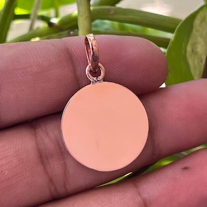 May include: A rose gold pendant with a circular design. The pendant is held up by a small loop at the top. The background features green leaves and vines.