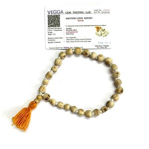 May include: A beaded necklace made of light brown, round beads with an orange tassel. The necklace is displayed with a white identification report from VEGGA GEMS TESTING LAB. The report details the necklace's specifications.