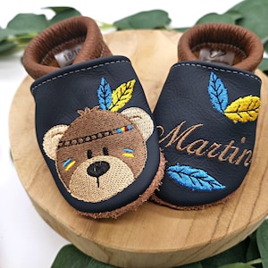 Könnte beinhalten: Ein Paar blaue Leder-Babymoccasins mit braunem Lederbesatz. Ein Moccasin zeigt einen gestickten Bären mit einem bunten Kopfschmuck und der andere Moccasin hat den Namen "Martin" gestickt.