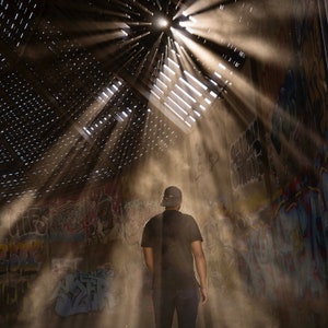 May include: A person wearing a black t-shirt and a black baseball cap stands in a dusty, graffiti-covered room.  Sunlight streams through a hole in the ceiling, casting beams of light across the room.