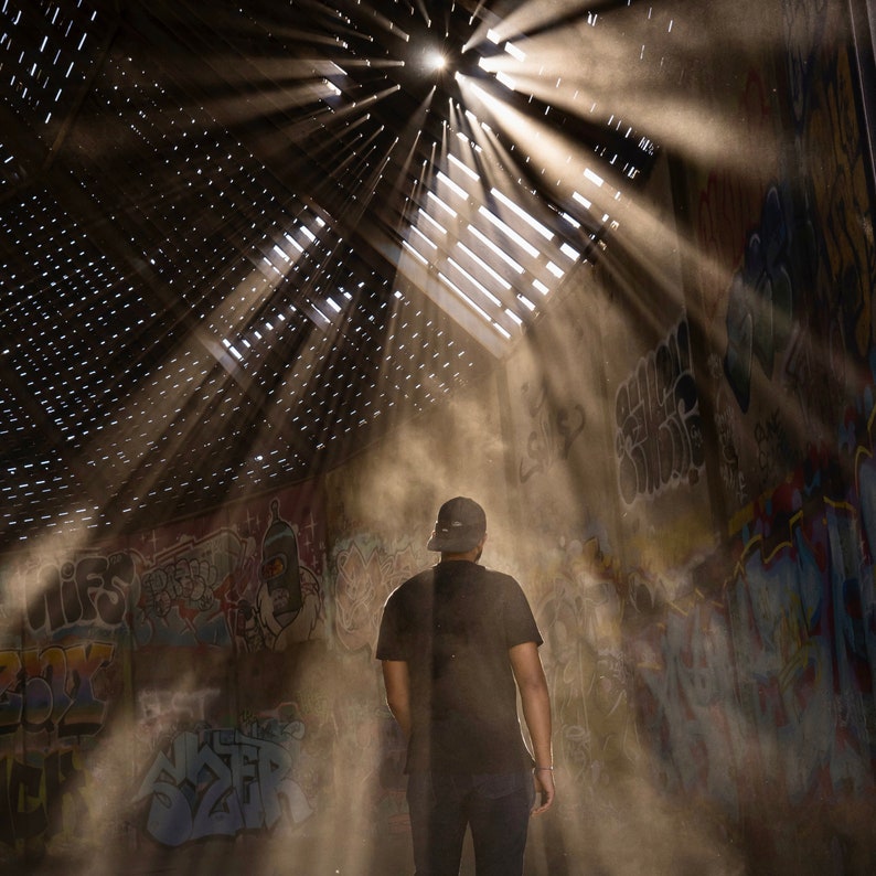 May include: A person wearing a black t-shirt and a black baseball cap stands in a dusty, graffiti-covered room.  Sunlight streams through a hole in the ceiling, casting beams of light across the room.